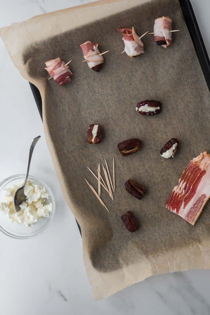 baking pan lined with parchment paper, with dates, goat cheese, bacon slices and toothpicks on top