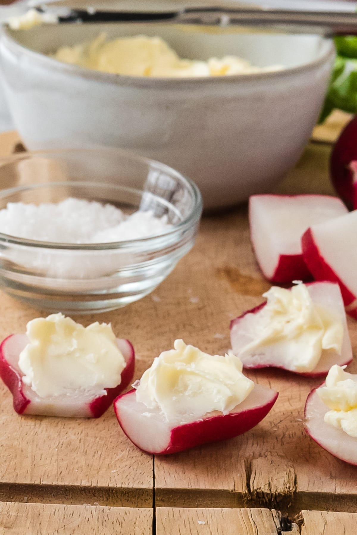 radishes cut in half, slathered with butter and sprinkled with salt on wood cutting board, with bowls of butter and salt in background