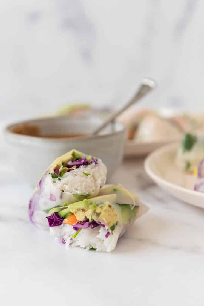 spring roll cut in half and stacked on top of each other, with plate and bowl of peanut sauce in background