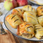 A plate of sausage rolls on a wooden table with apples in the background